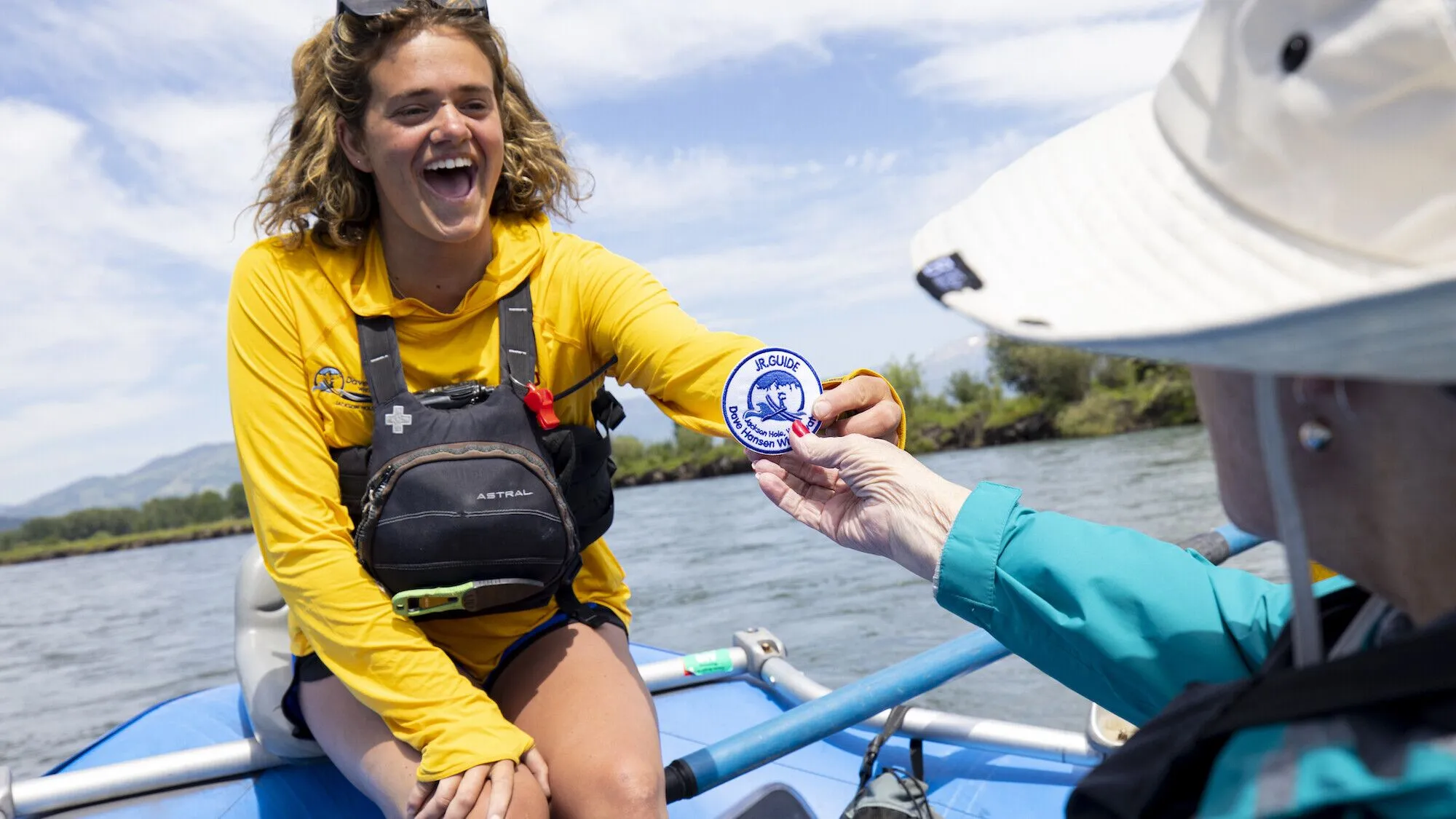 A cheerful scenic rafting guide in a yellow shirt hands an elderly woman a JR Guide patch.