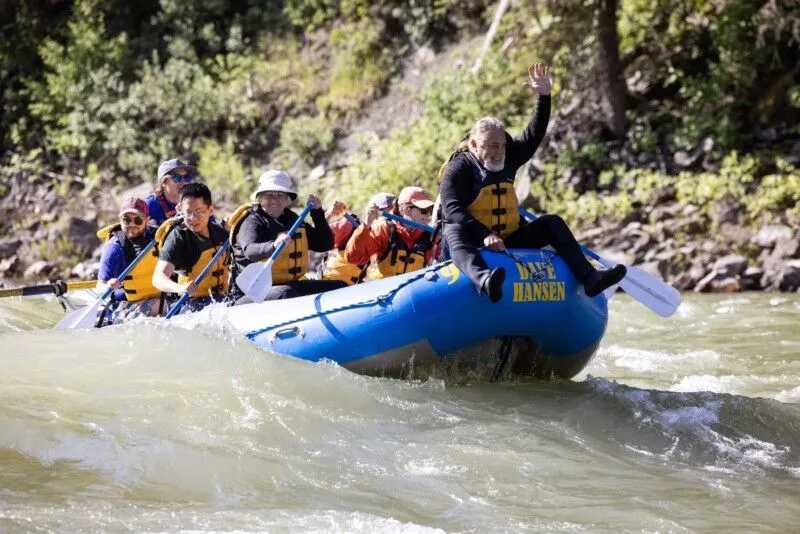 A man with a white beard rides on the front of a blue Dave Hansen Whitewater raft as they paddle through a whitewater rapid.