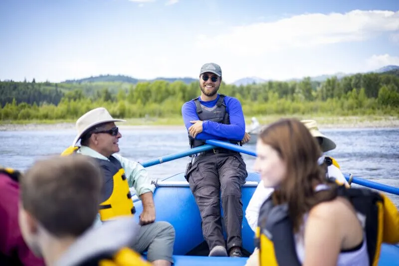 A scenic raft guide relaxes on the edge of a blue raft on a sunny day.
