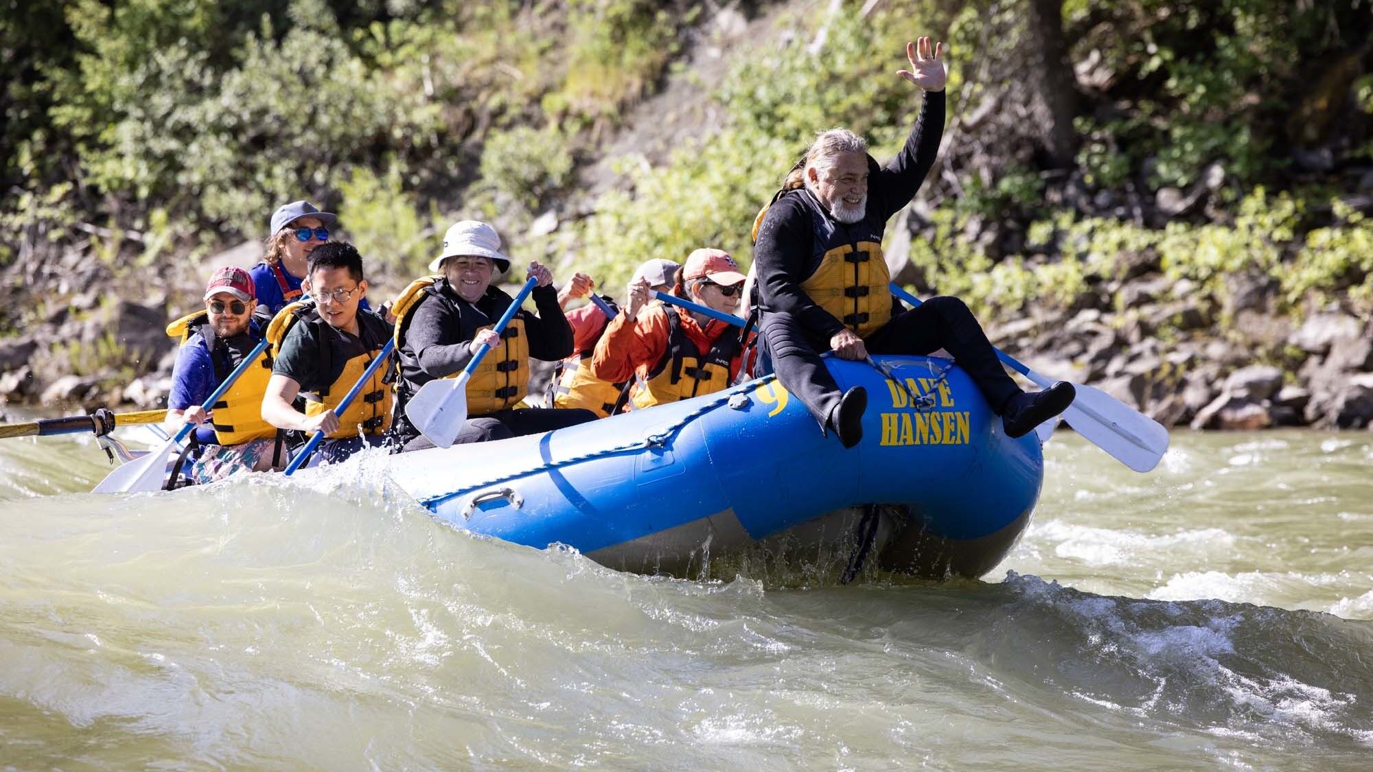 A man with a white beard rides on the front of a blue Dave Hansen Whitewater raft as they paddle through a whitewater rapid.