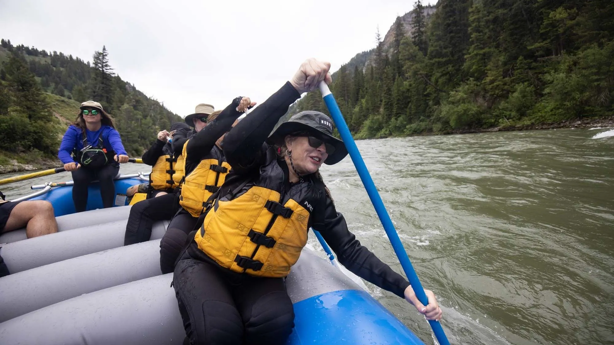 An older woman helps paddle a blue raft down the Snake River on a rainy day in Jackson Hole.