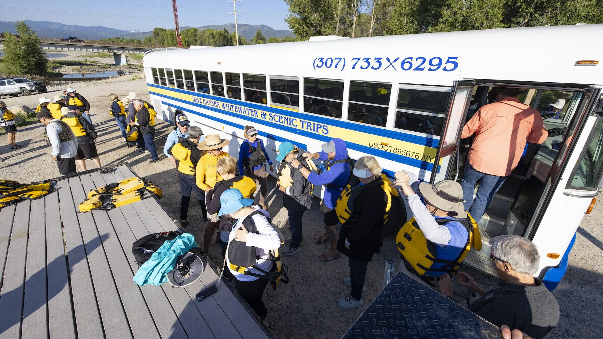 A group of rafting clients putting on yellow lifejackets next to the blue, white and yellow Dave Hansen Whitewater & Scenic Trips bus.