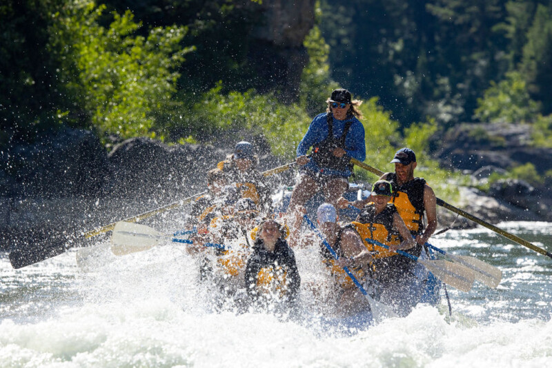 A group of children and adults splash a blue Dave Hansen Whitewater raft through whitewater rapids on the Snake River.