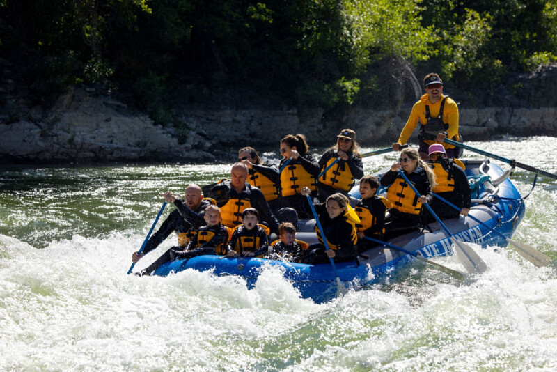 A group of children and adults paddling a blue Dave Hansen Whitewater raft through whitewater rapids on the Snake River.