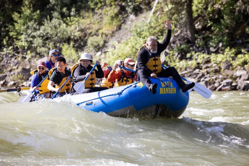 A man with a white beard rides on the front of a blue Dave Hansen Whitewater raft as they paddle through a whitewater rapid.
