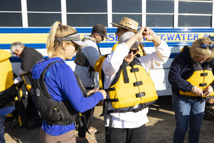 A rafting guide helps an elderly woman put on a yellow lifejacket.