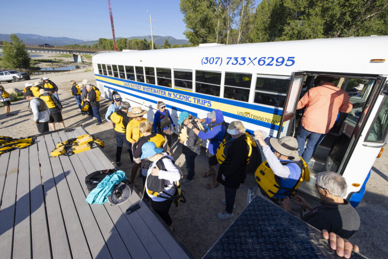 A group of rafting clients putting on yellow lifejackets next to the blue, white and yellow Dave Hansen Whitewater & Scenic Trips bus.