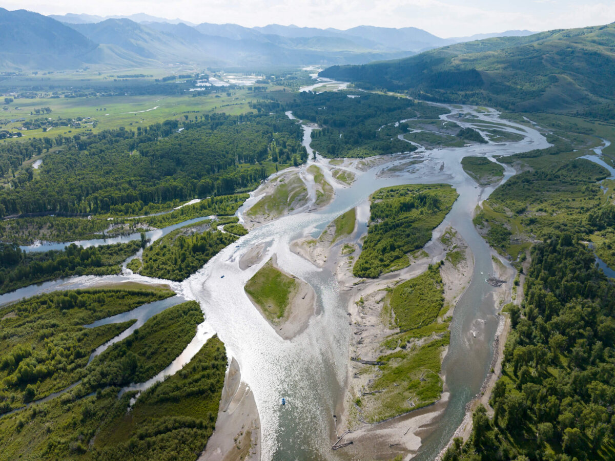 Sweeping high-altitude aerial drone photo of the Snake River's multiple braided channels spreading across the wide Jackson Hole valley floor, with forested hills, farmland, and hazy mountains in the distance.