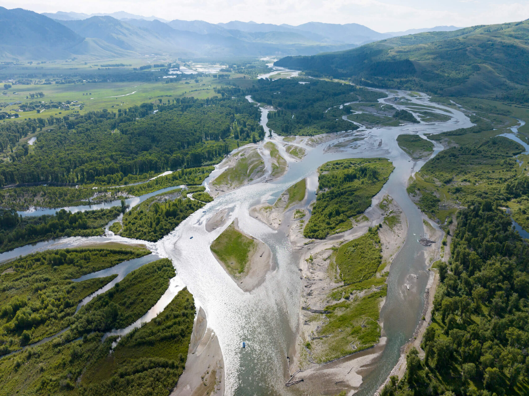 Sweeping high-altitude aerial drone photo of the Snake River's multiple braided channels spreading across the wide Jackson Hole valley floor, with forested hills, farmland, and hazy mountains in the distance.