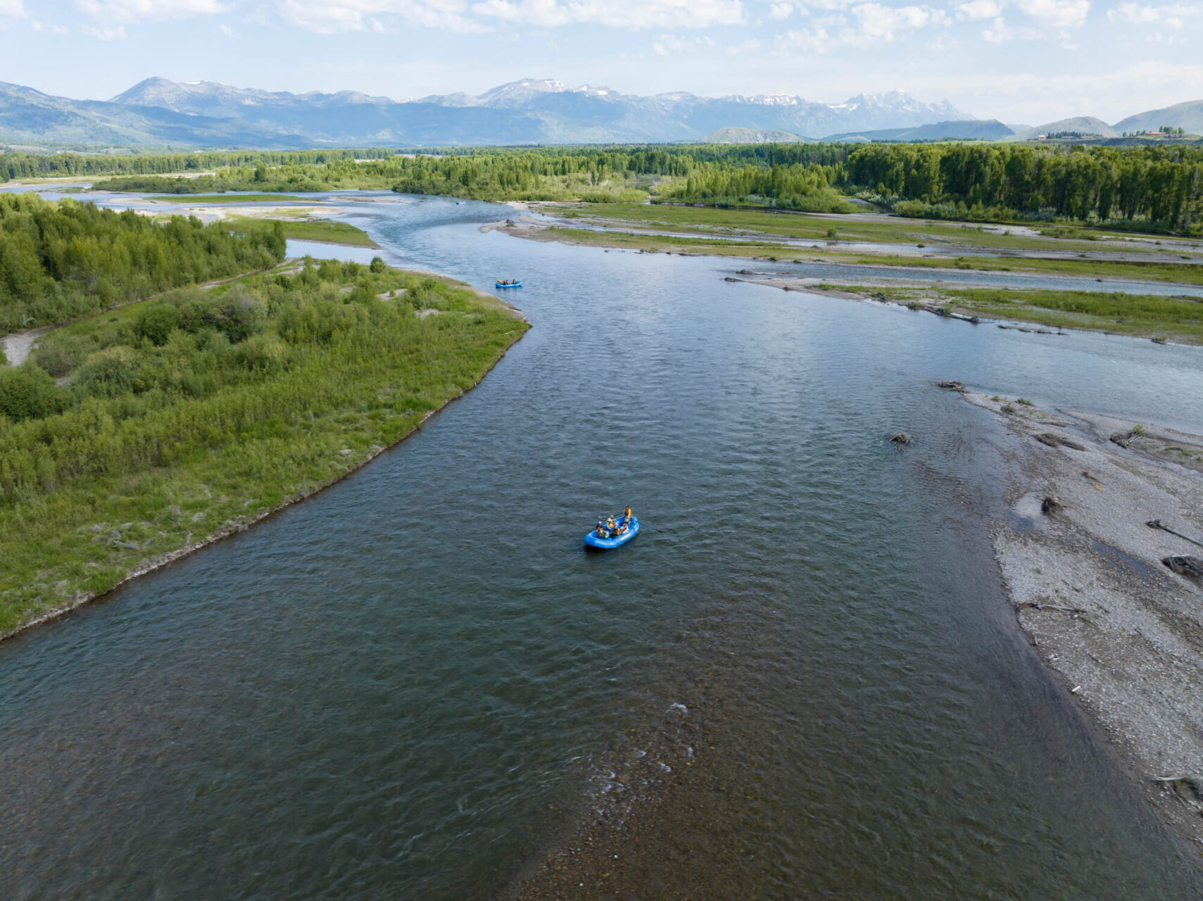Aerial drone photo of two Dave Hansen River Trips rafts floating in formation down a wide, calm stretch of the Snake River, surrounded by green riverbank vegetation and snow-capped mountains in the distance.