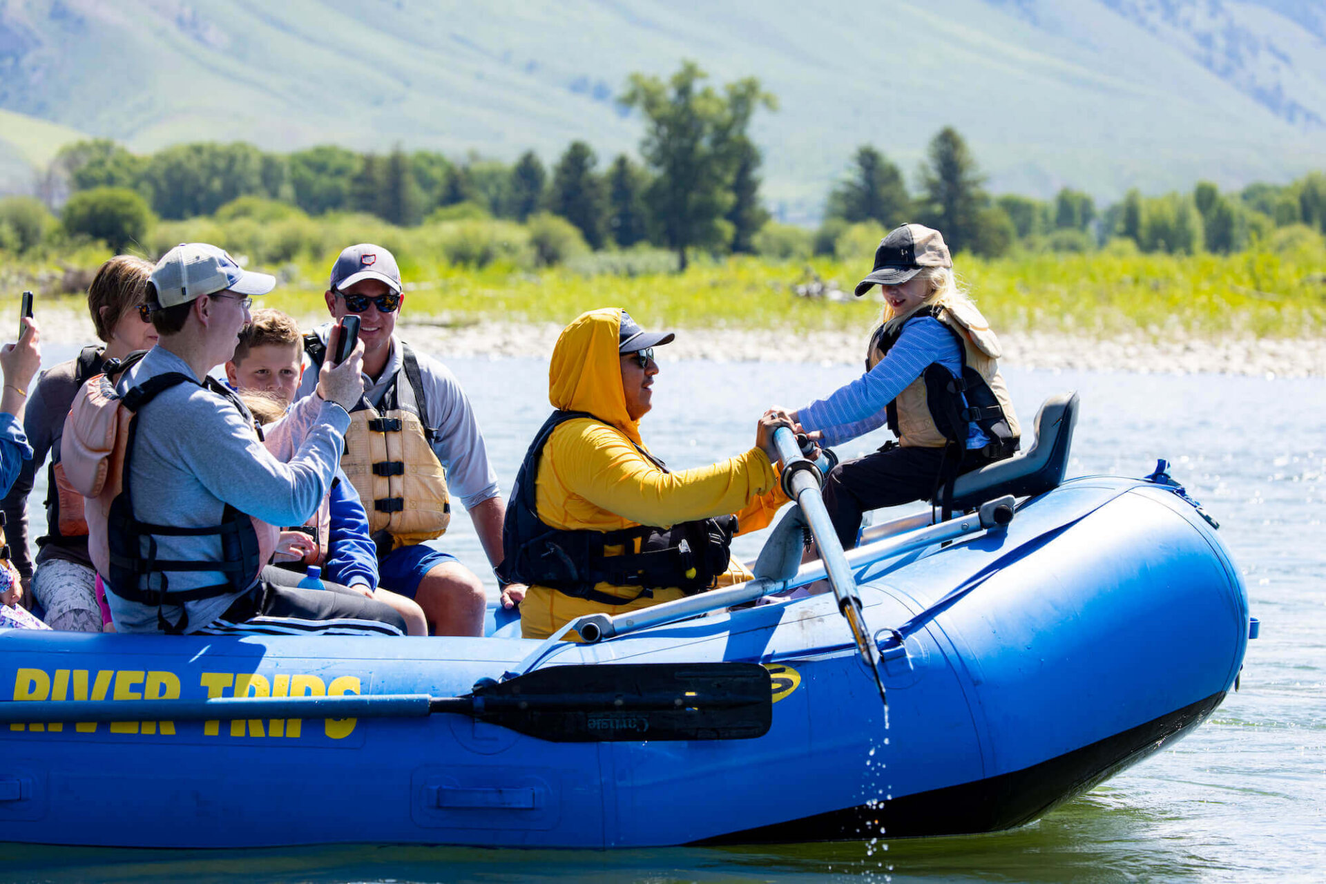 A family with children boarding a Dave Hansen River Trips raft on a calm river stretch in Jackson Hole, Wyoming, with green meadows and mountains visible in the background.