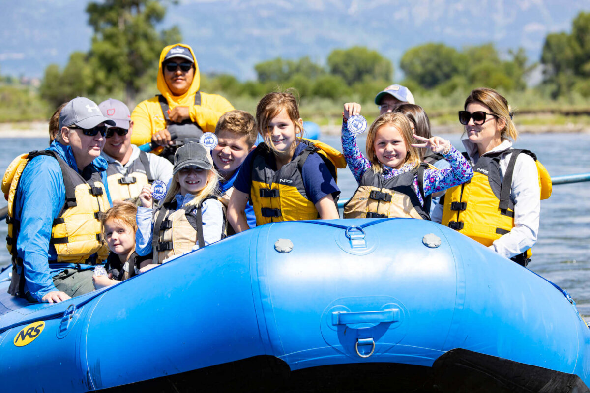 A family of adults and young children smiling and holding up Dave Hansen River Trips patches at the end of a scenic float trip, with the blue raft and green trees behind them.