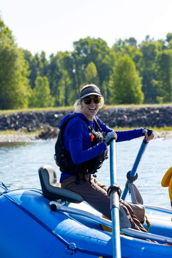 A smiling female Dave Hansen River Trips guide in a blue jacket and visor rows a raft on a sunny stretch of the Snake River, with a green riverbank and bright blue sky behind her.