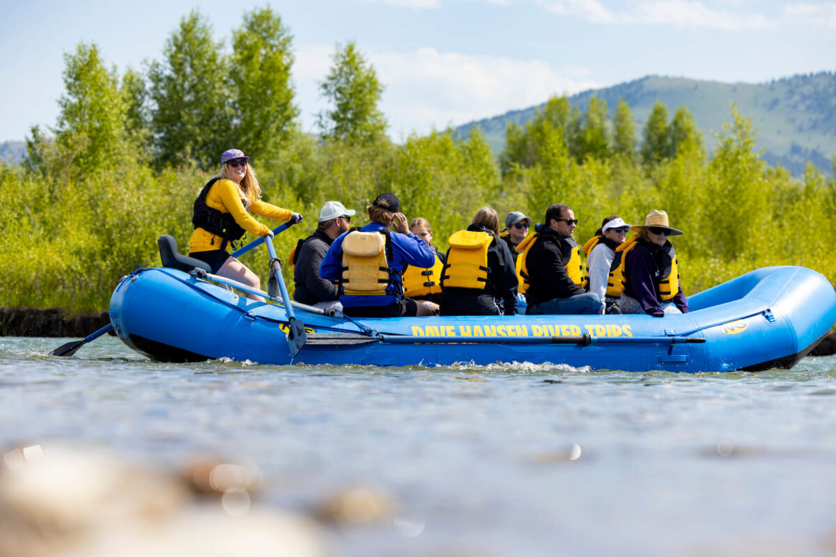 A smiling female Dave Hansen River Trips guide in a yellow shirt stands at the oars of a raft full of adult guests in yellow life jackets on a calm, sunny stretch of the Snake River.