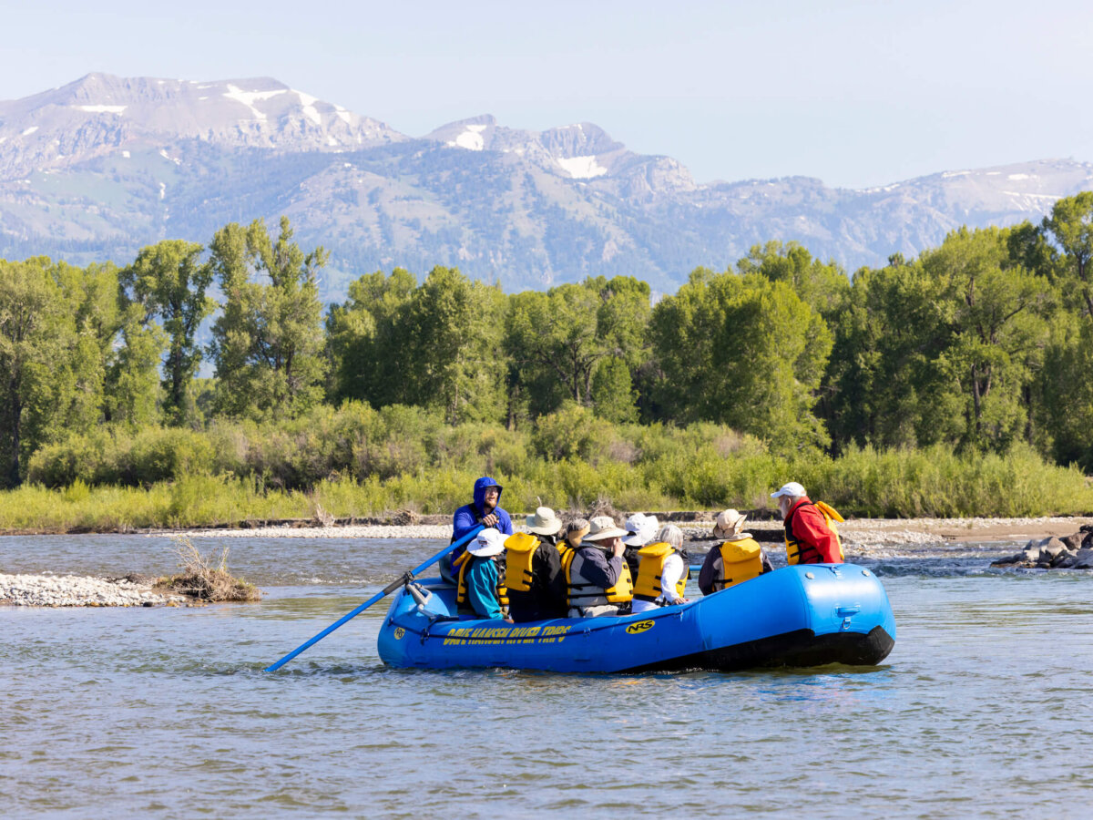 A Dave Hansen River Trips raft with a group of adults in yellow life jackets floats past a rocky riverbank on the Snake River, with snow-dusted mountain peaks visible behind green cottonwood trees.
