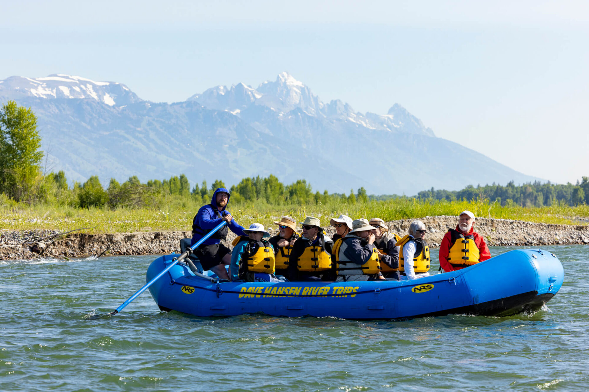 A Dave Hansen River Trips guide in a blue hoodie rows a full raft of guests down the Snake River, with the iconic jagged Grand Teton peaks rising behind a green tree line.