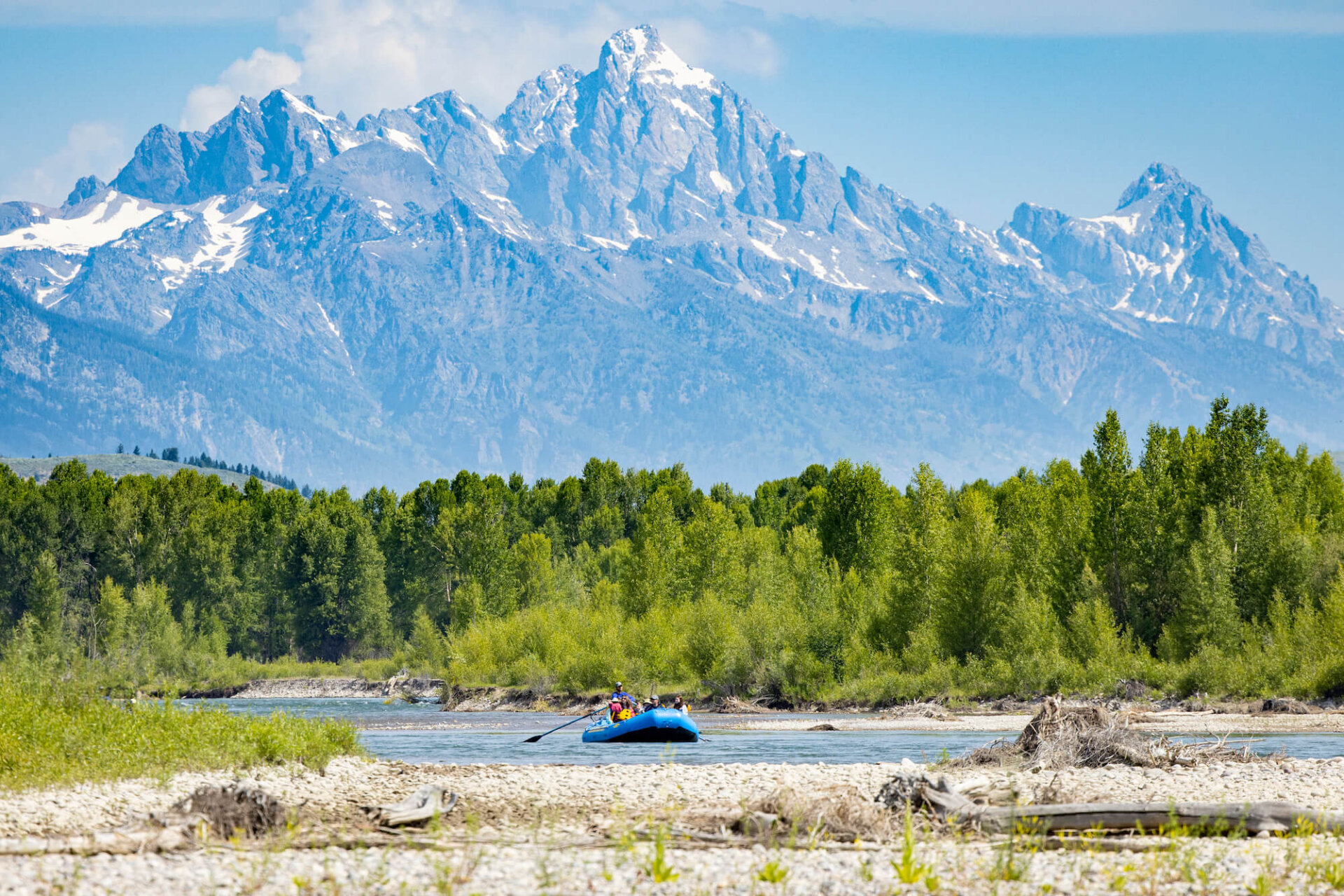 A lone Dave Hansen River Trips raft glides along the Snake River with the dramatic snow-covered Grand Teton peaks filling the skyline behind lush green cottonwood and aspen trees on a clear summer day.