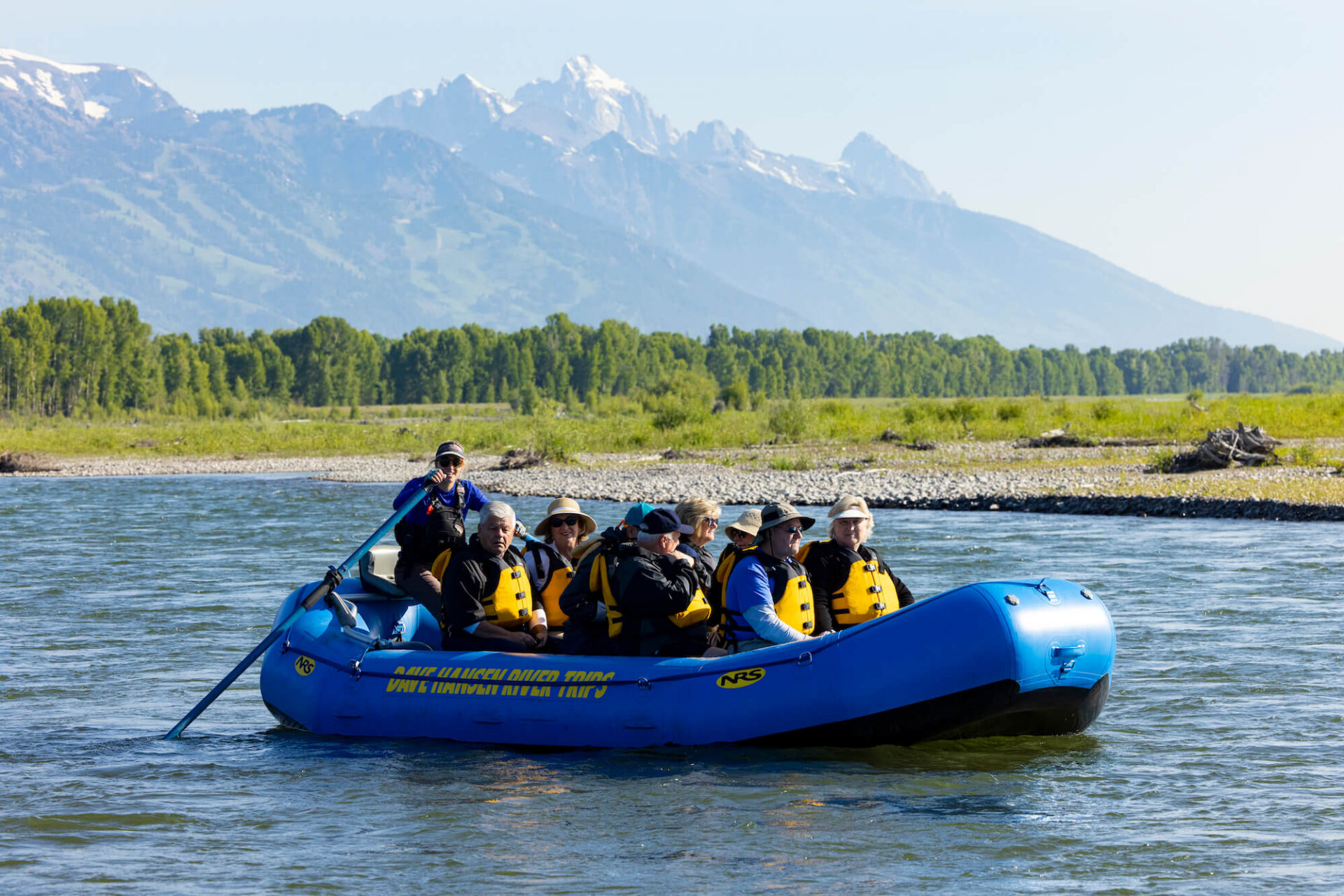 A Dave Hansen River Trips raft carrying a group of adults on a calm float down the Snake River, with the snow-dusted Grand Teton peaks towering behind a green tree line.