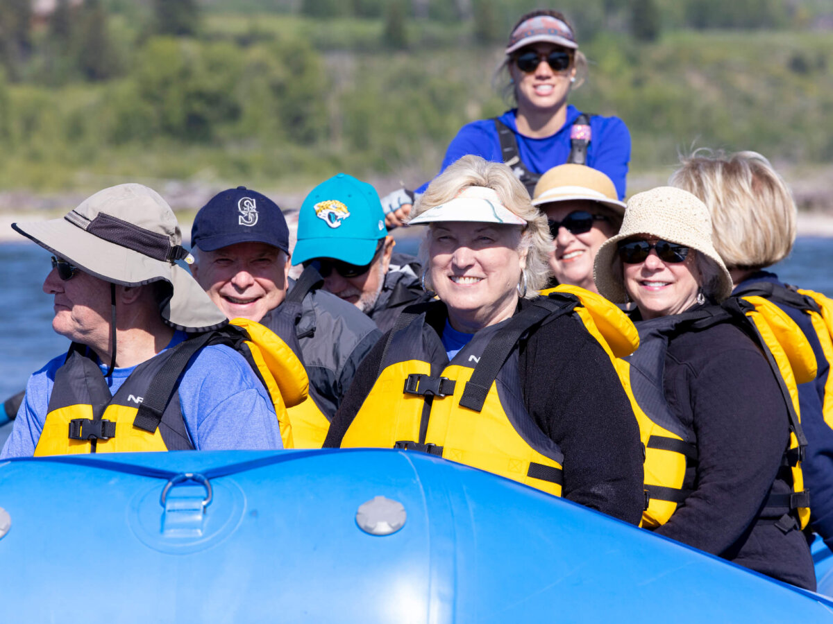 A group of smiling adults seated on a Dave Hansen River Trips raft look toward the camera on a sunny scenic float, with a female guide standing at the stern and green riverbank trees behind them.
