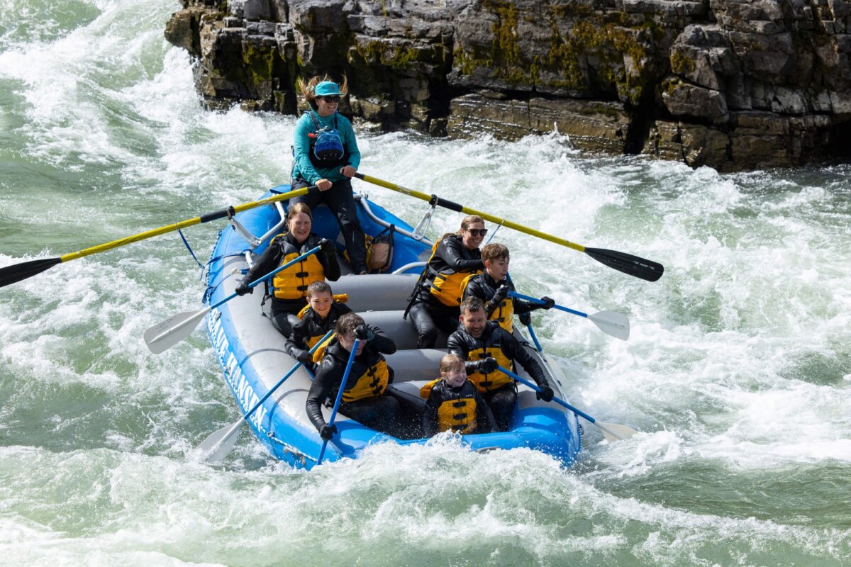 A Dave Hansen guide in a teal jacket rows a raft full of smiling families through a lively rapid in the Snake River Canyon, with rocky walls and green vegetation on the riverbank behind them.