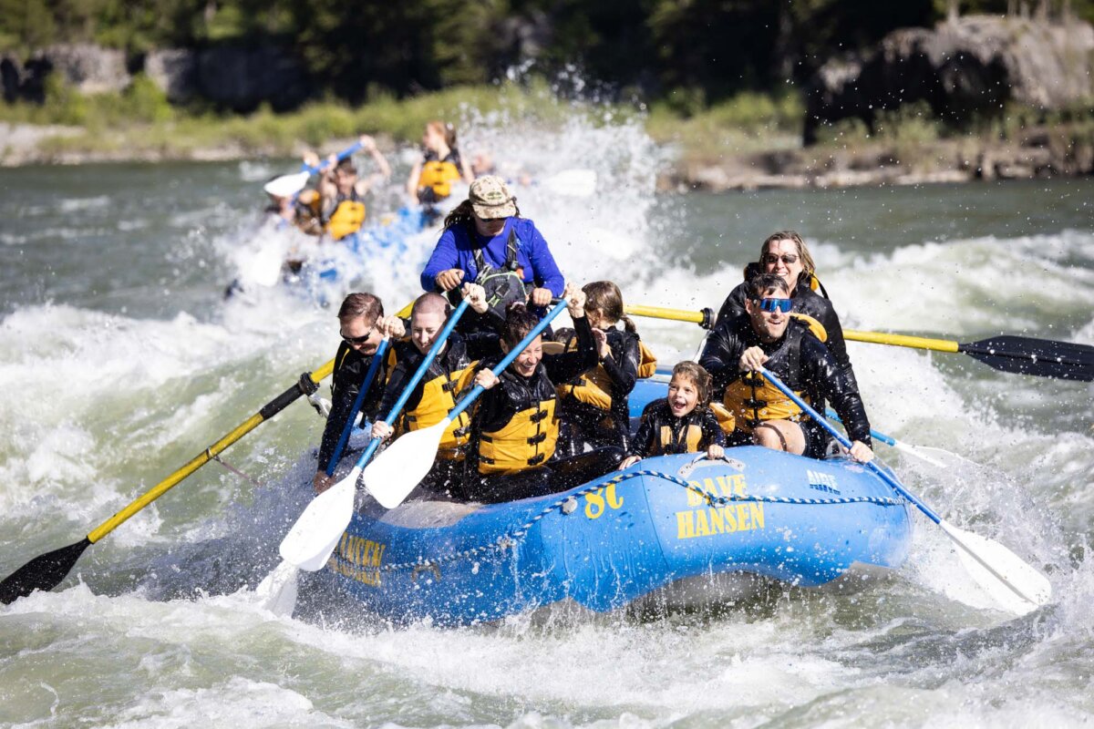 Two Dave Hansen whitewater rafts navigating rapids on a sunny river, with smiling families and children in yellow life jackets paddling together.