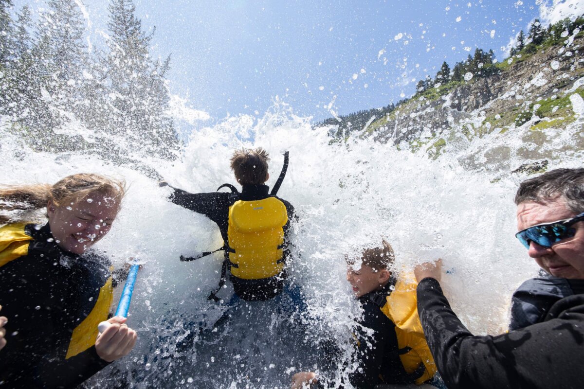 A family on a Dave Hansen raft braces as a massive wave crashes over them in the Snake River Canyon, water exploding in all directions against a bright blue sky and forested canyon walls.