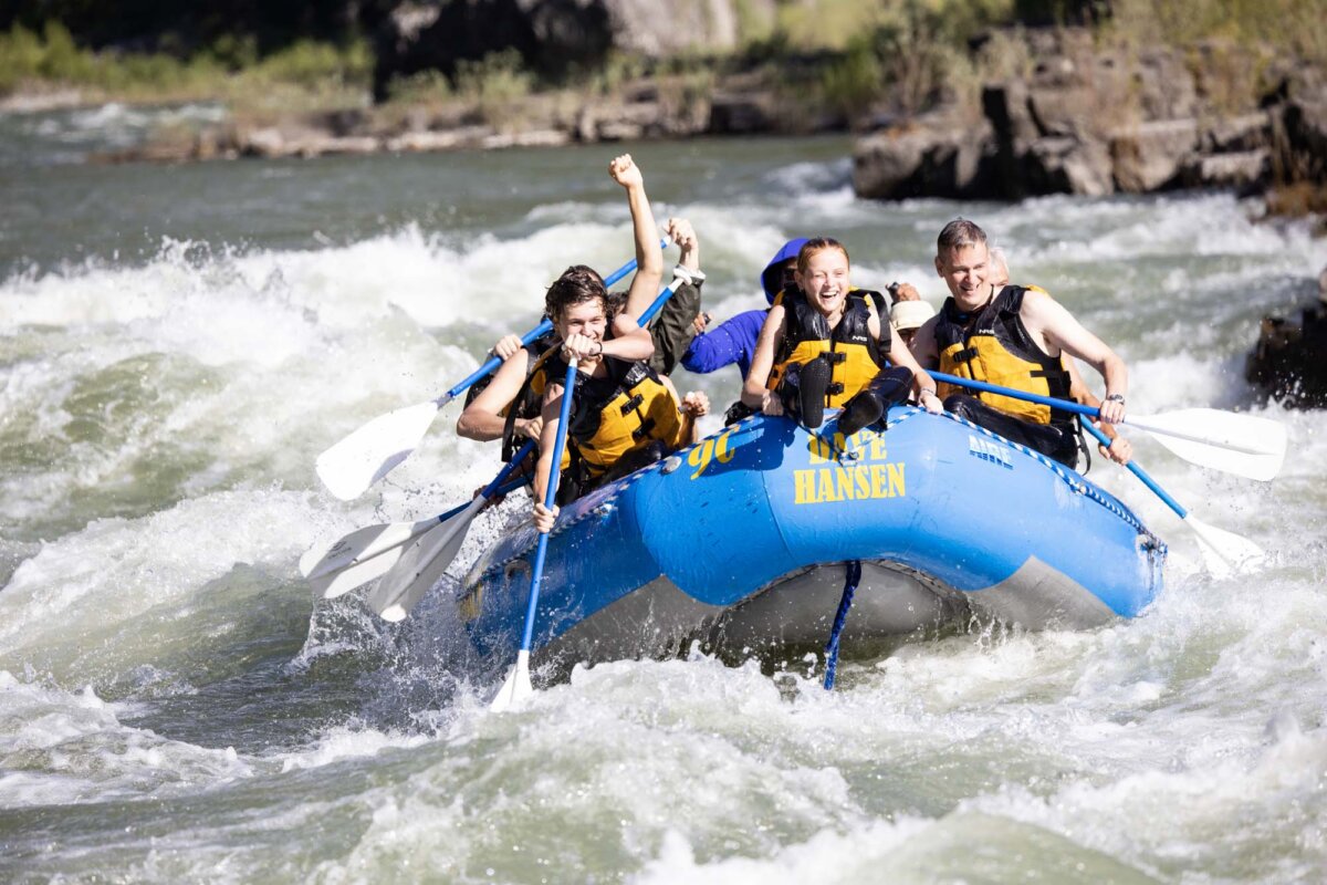 A group of adults cheering and raising their fists in celebration while paddling a Dave Hansen blue raft through whitewater rapids with rocky canyon walls in the background.