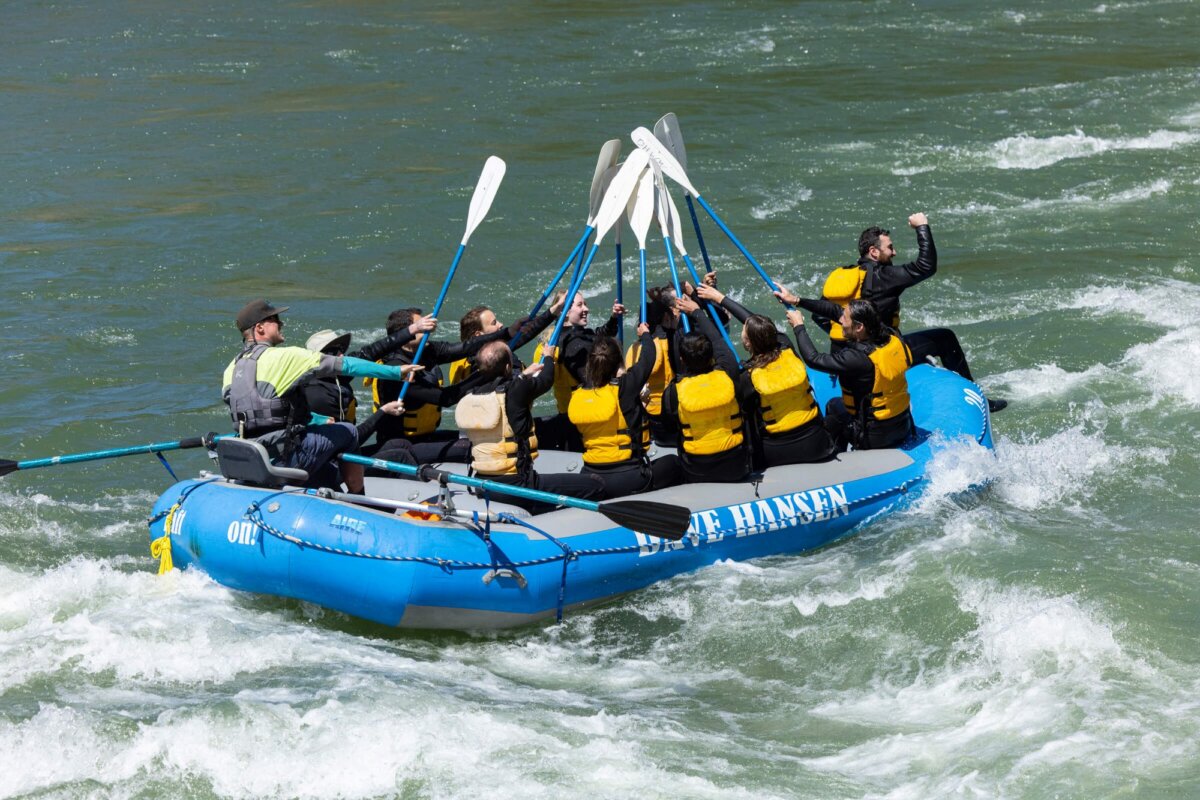 A full Dave Hansen raft of guests raise all their paddles together in a celebratory salute after conquering a whitewater rapid, with one rider standing and cheering arms raised.