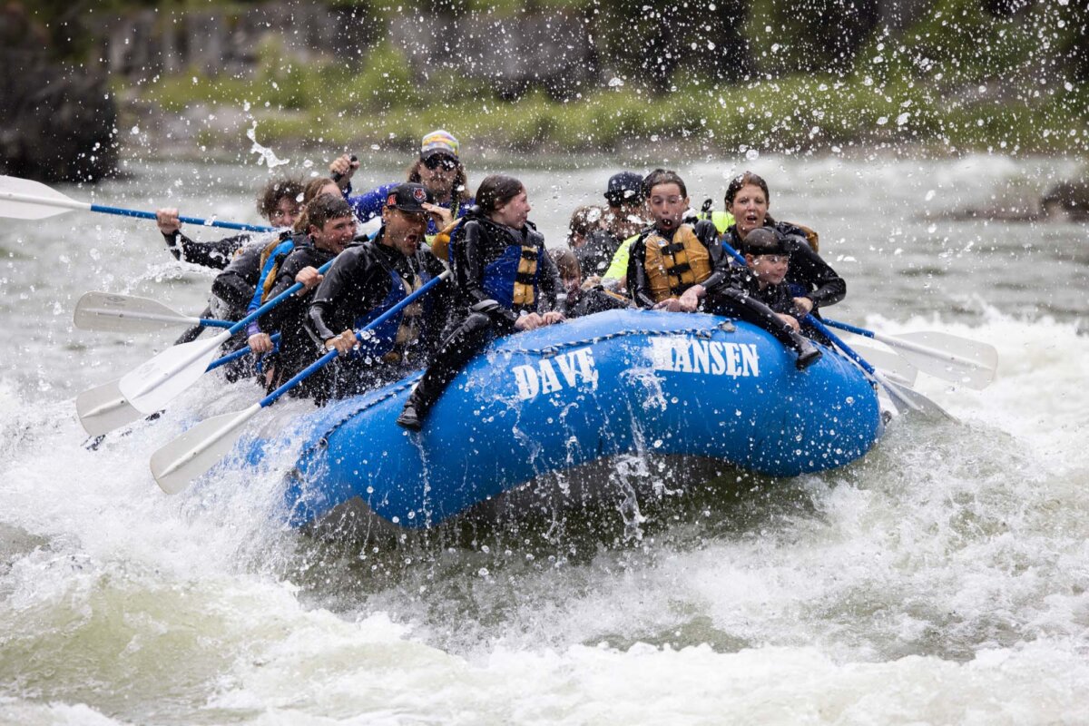 A large group of young rafters paddling through churning whitewater rapids on a Dave Hansen blue inflatable raft, paddles raised and faces showing excitement and determination.