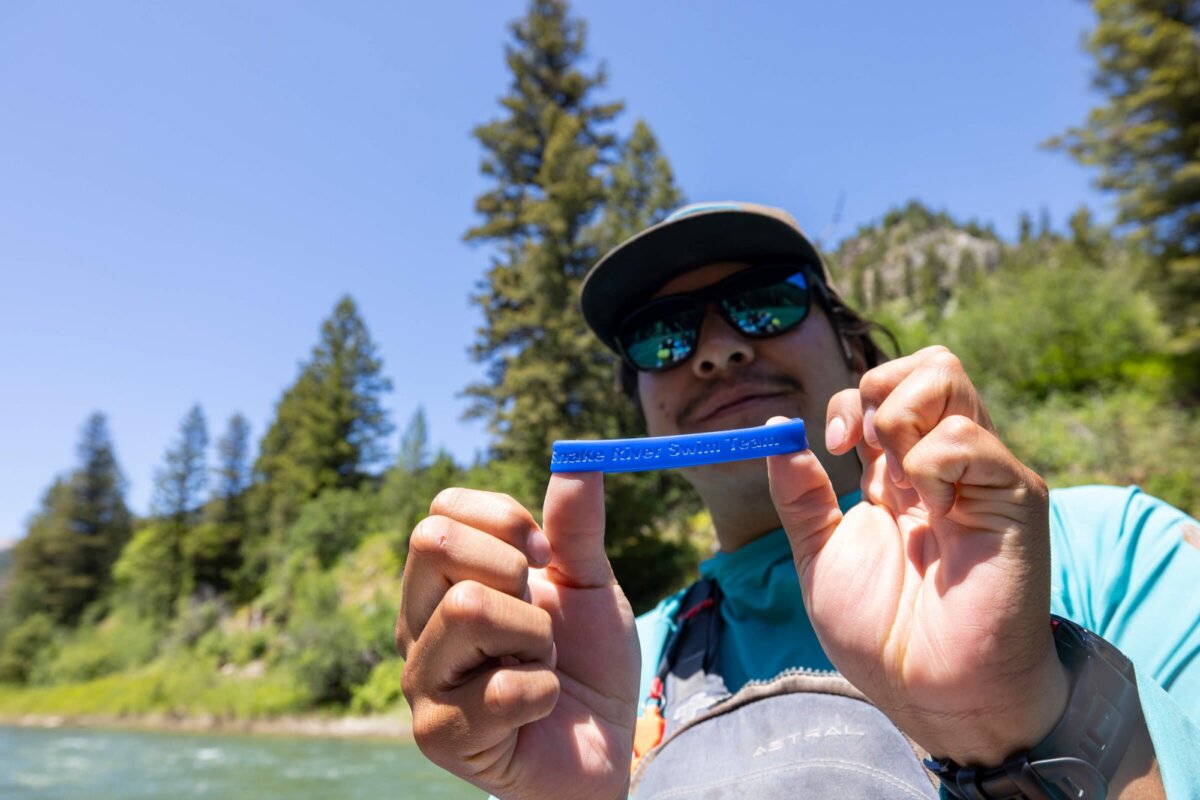 A Dave Hansen River Trips guide holds up a blue Snake River Swim Team rubber bracelet toward the camera with a grin, the Snake River Canyon pine forest visible behind them on a clear day.