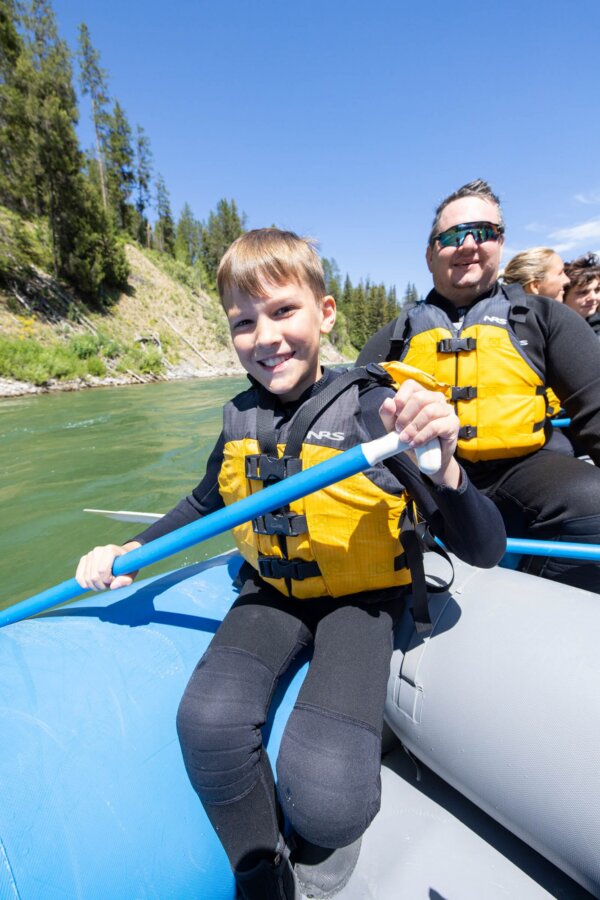 A young boy smiles confidently at the camera while holding a paddle on a Dave Hansen raft in the Snake River Canyon, with his dad and other guests visible behind him on a sunny day.