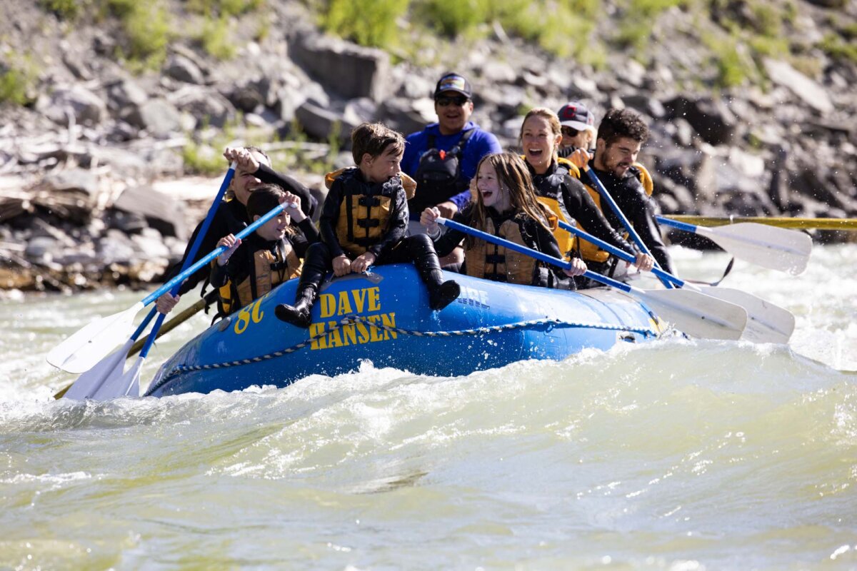 Young children perched on the bow of a Dave Hansen raft, laughing and holding on as the boat powers through a whitewater rapid with rocky canyon walls in the background.