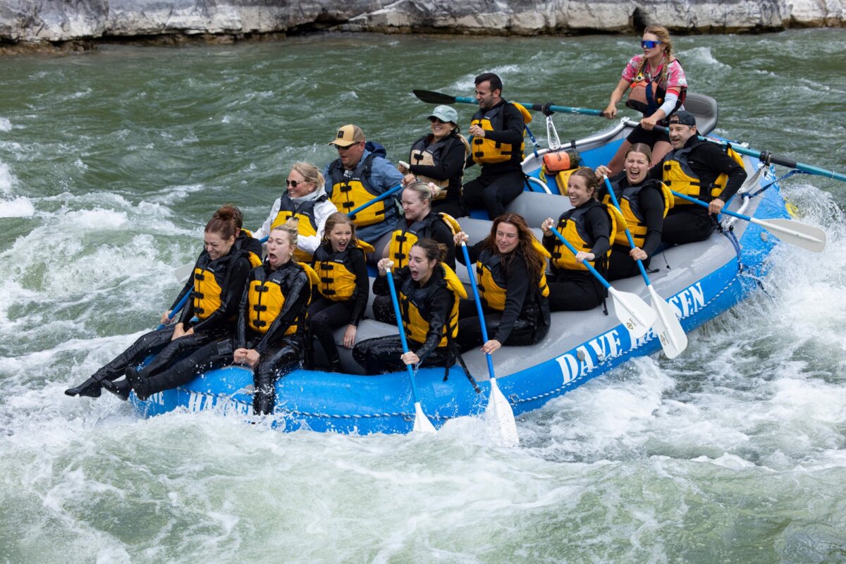 A large mixed group of adults screaming and paddling through churning whitewater rapids on a Dave Hansen raft in the Snake River Canyon, with grey limestone walls in the background on an overcast day.