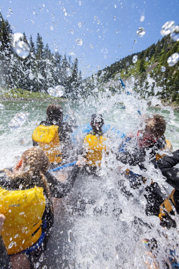 An explosive wall of whitewater engulfs rafters on a Dave Hansen raft as they hit a rapid in the Snake River Canyon, with droplets suspended mid-air against a blue sky and pine-covered ridgeline.