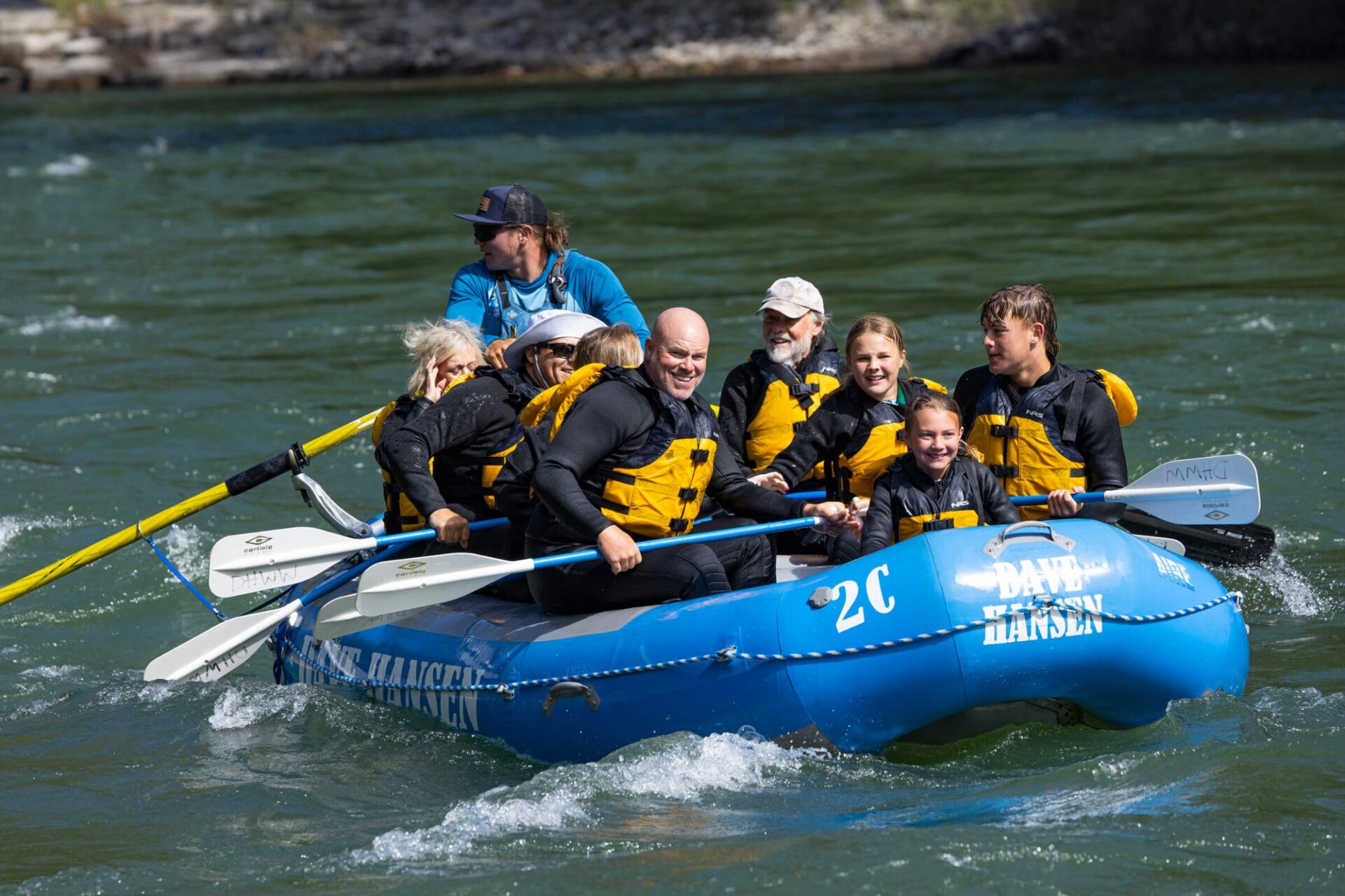 A multigenerational family — grandparents, parents, and kids — smiles broadly on a Dave Hansen raft navigating a calm stretch of the Snake River Canyon, with a guide steering from the back.