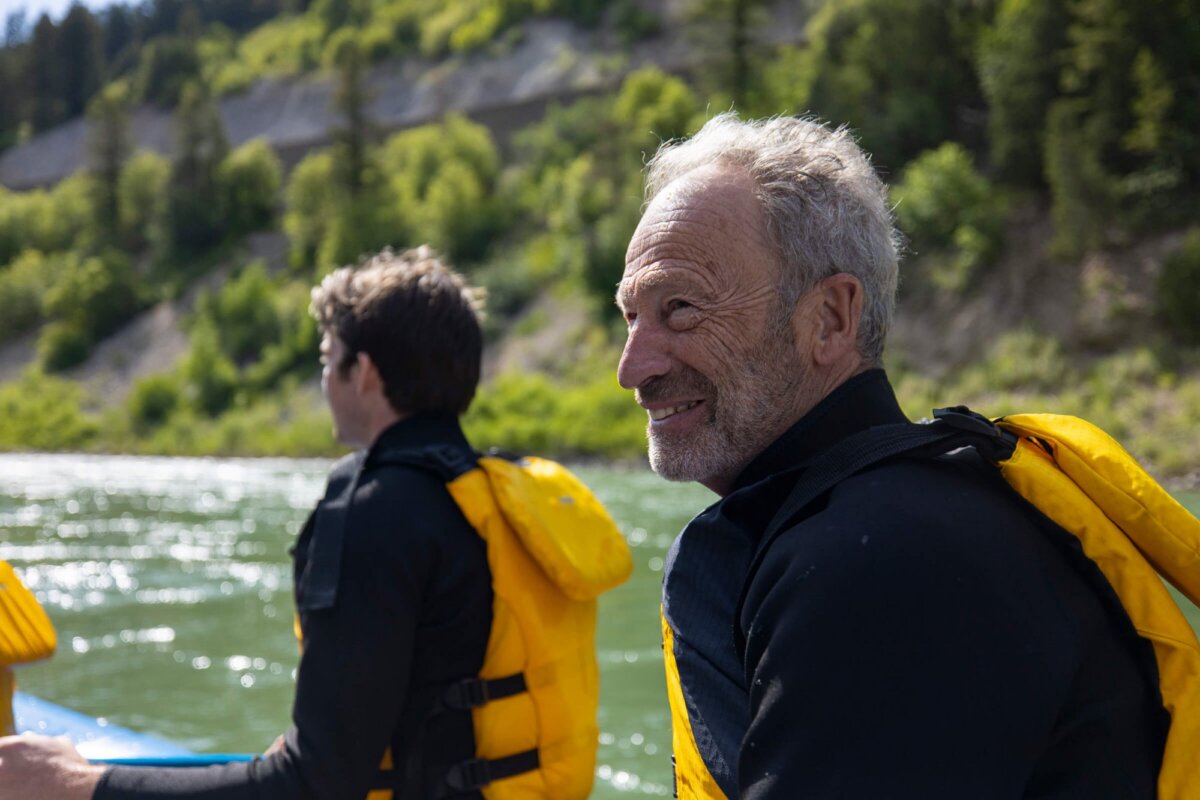 A grey-haired man in a yellow life jacket turns and smiles warmly at the camera from a Dave Hansen raft on the Snake River Canyon, with green willows and canyon cliffs visible behind him.