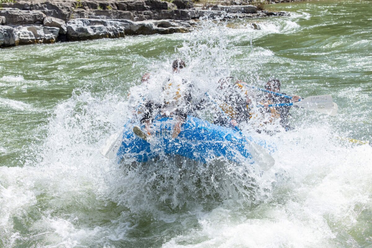 A Dave Hansen raft is almost completely swamped by a massive whitewater wave in the Snake River Canyon, with rafters' legs and paddles visible above the wall of white water.