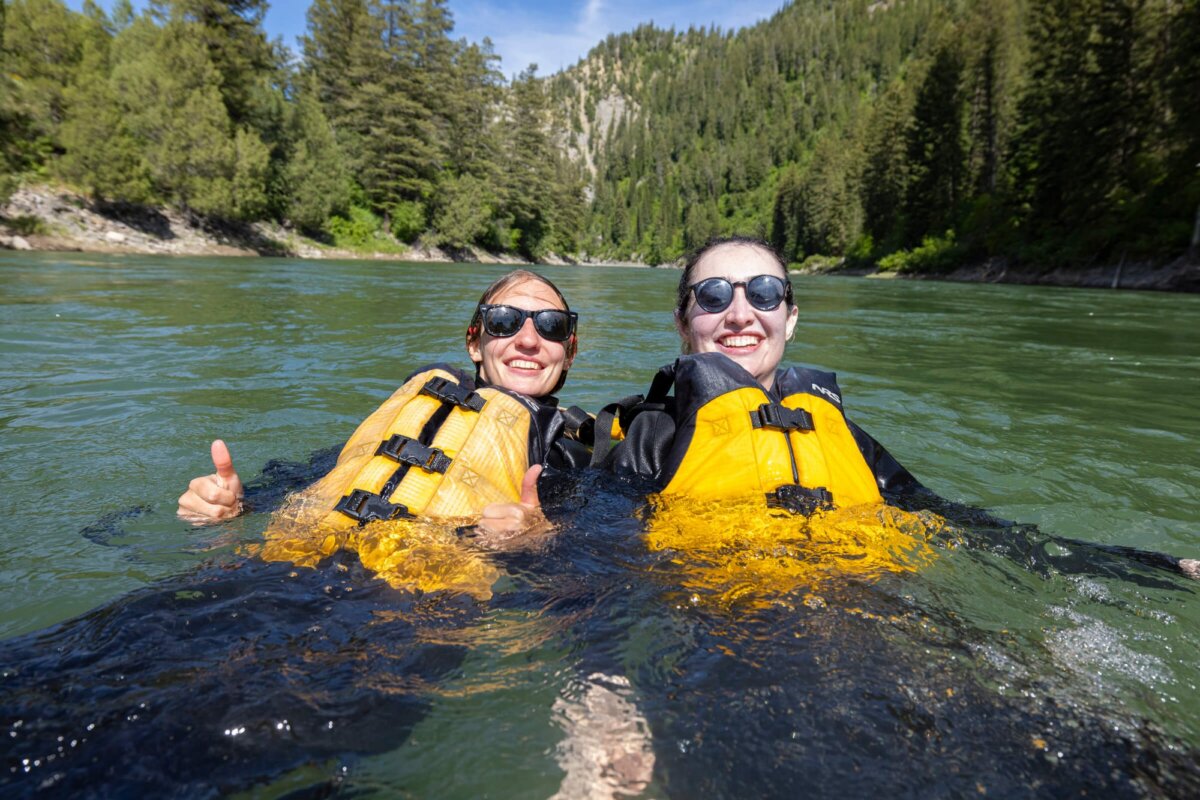 Two women in yellow life jackets smile and give a thumbs-up while floating in the calm green Snake River Canyon after going for a swim during a Dave Hansen whitewater rafting trip.