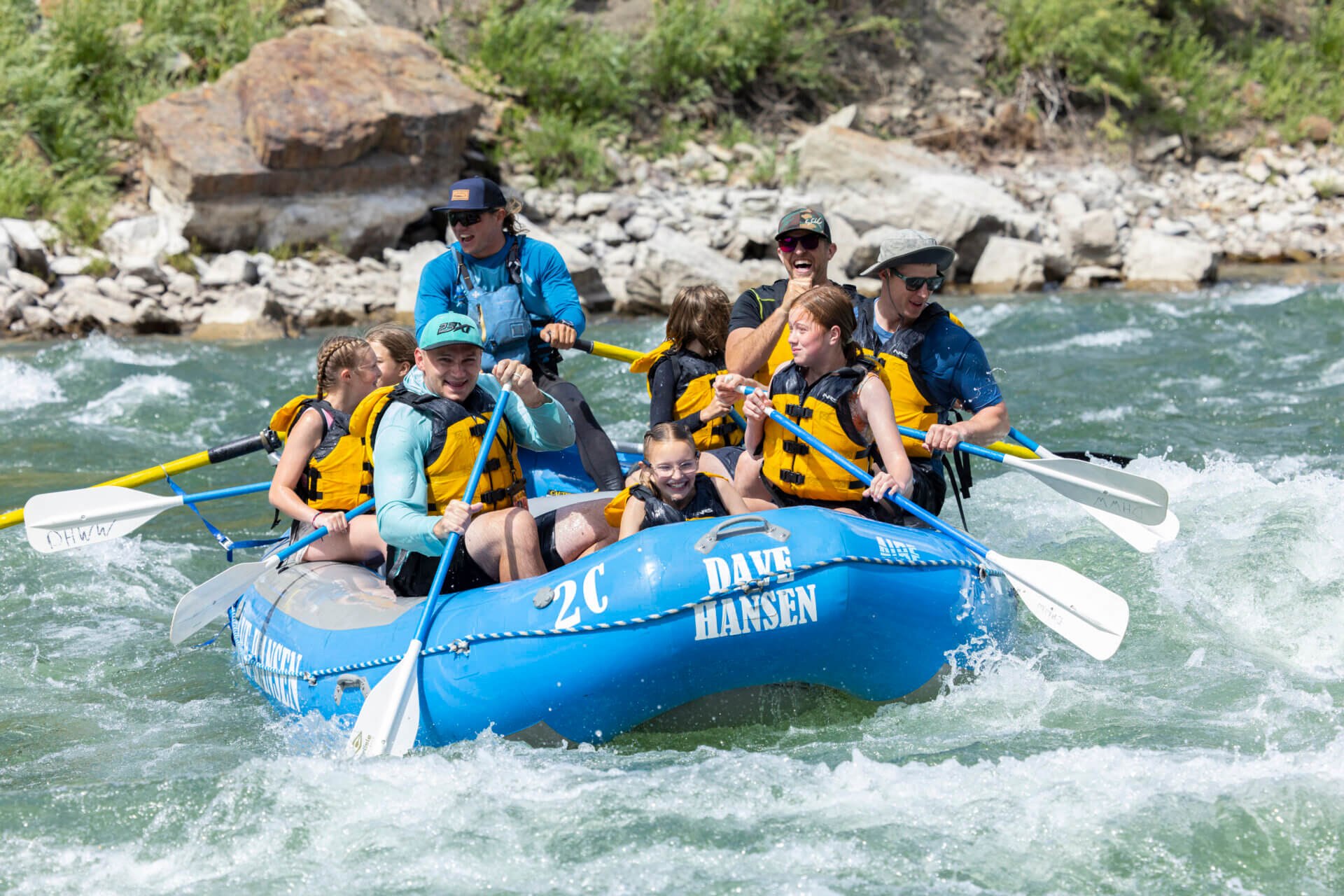 A group of happy families and children paddling together through rapids on a blue Dave Hansen Whitewater raft.