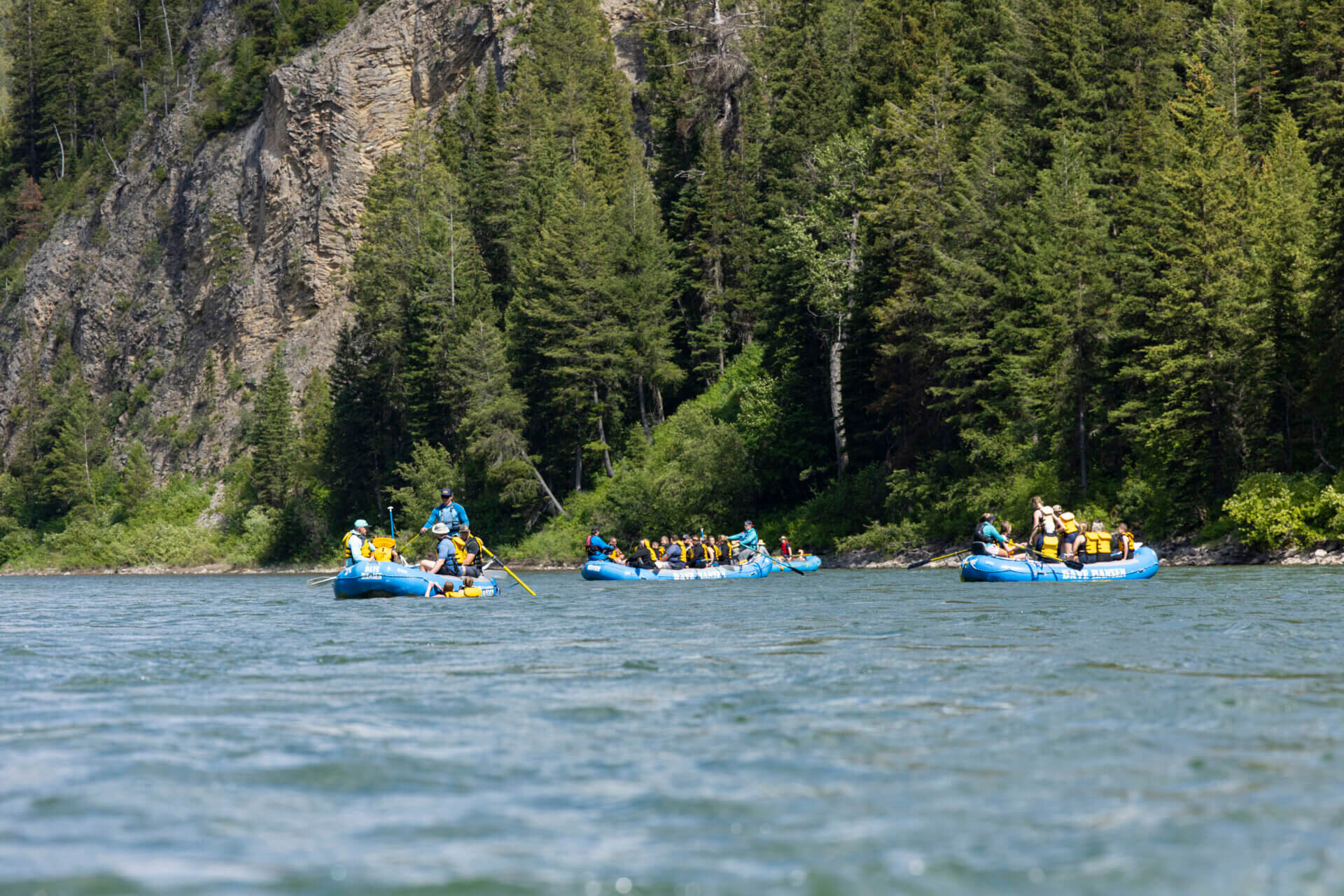 Multiple blue Dave Hansen Whitewater rafts floating down a calm river with people swimming and canyon scenery.
